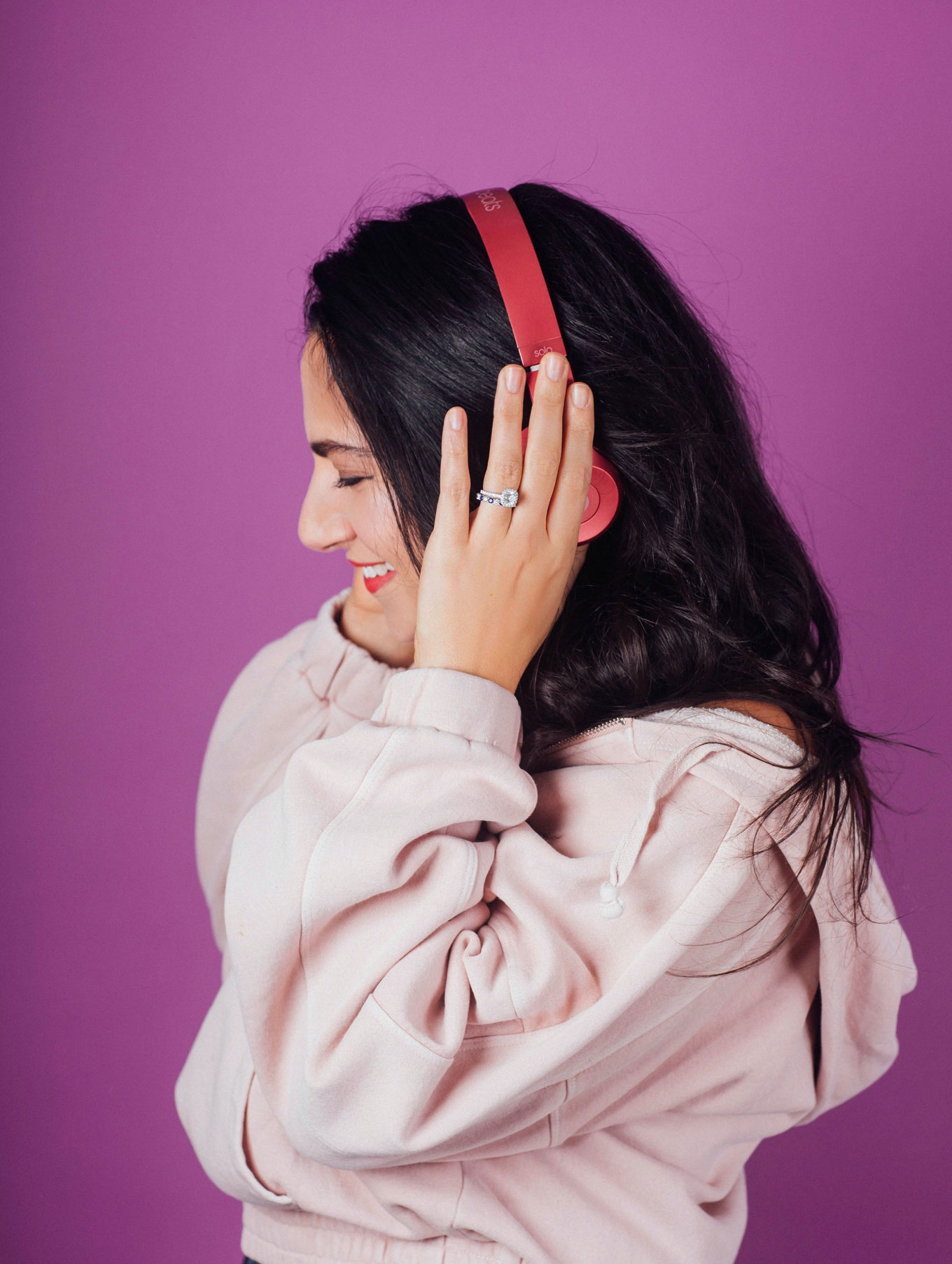 A young female listening to music in red headphones wearing a light pink hoodie and red lipstick
