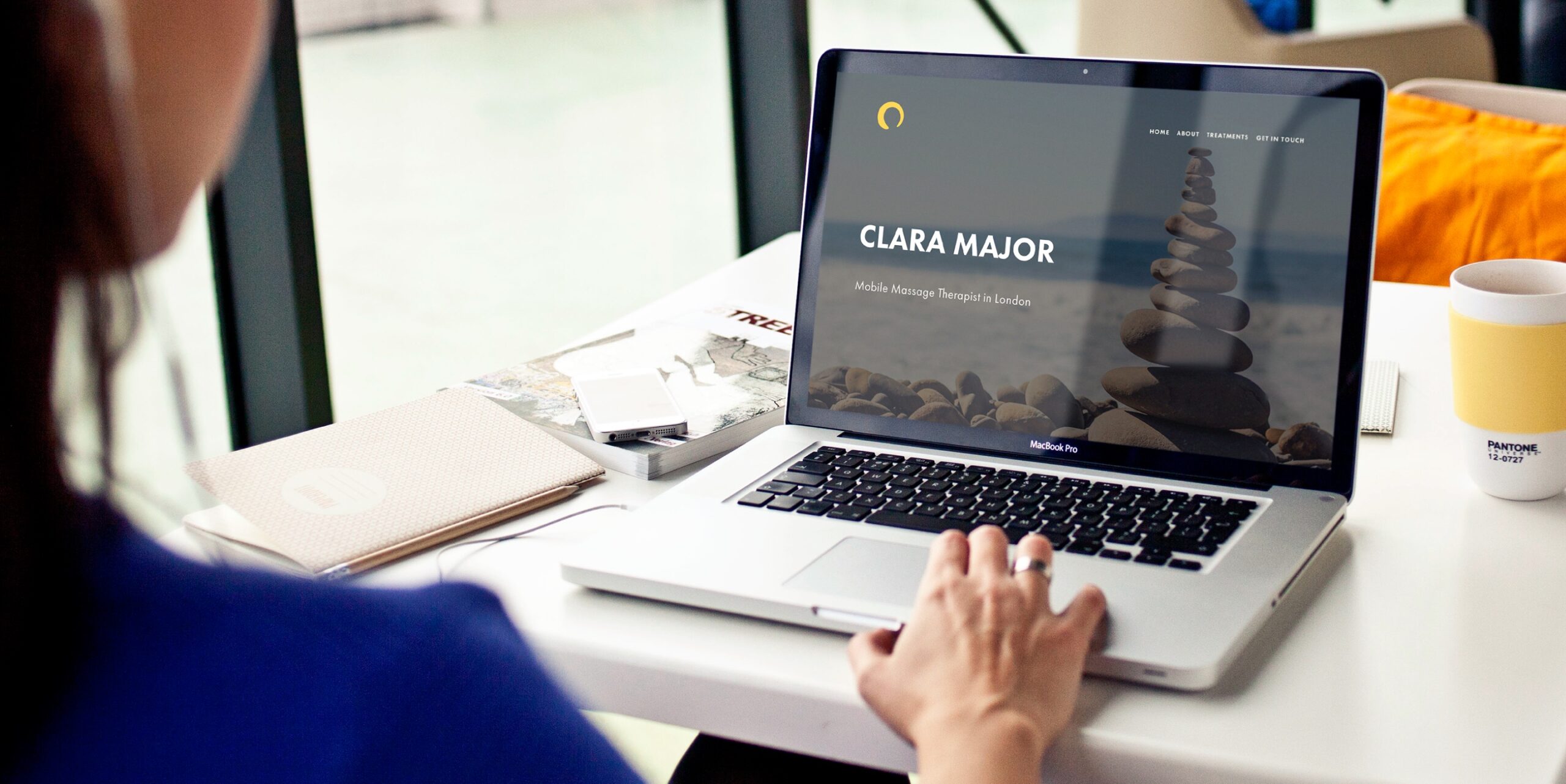 A woman sitting by a desk and looking at her macbook, seeing Clara Major' website on the screen